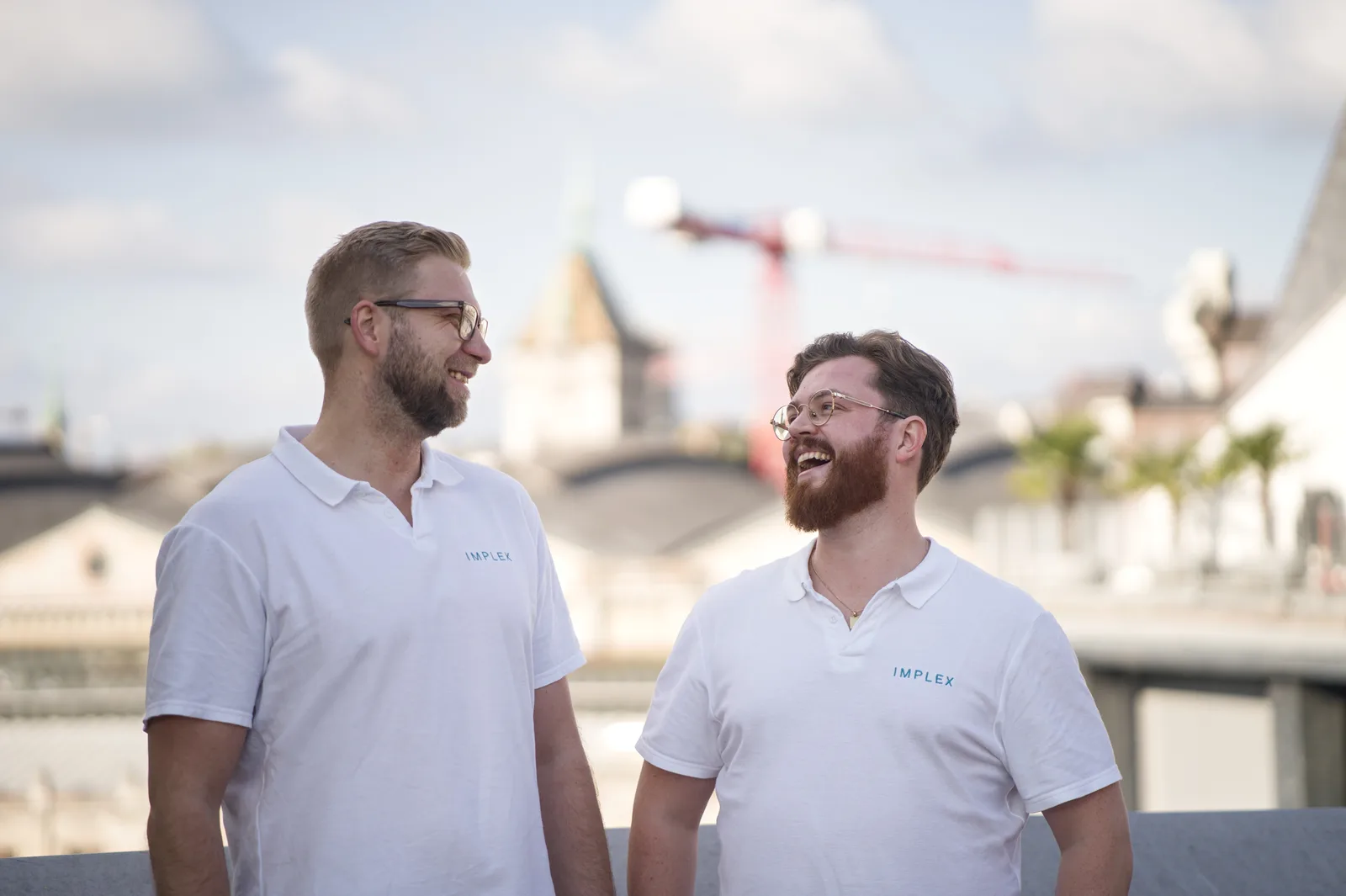 Nils Bohrsen und Torben Schulthoff, Gründer von Implex, lachen auf einer Dachterrasse mit Hamburger Skyline